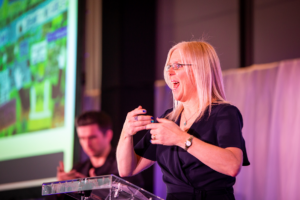 Rebecca Mansell, a white woman with long blonde hair and glasses stands behind a clear podium, giving a presentation. She is wearing a black outfit and blue nail polish. In the background, another person is slightly out of focus. A large screen with colourful visuals and text is partially visible on the left.