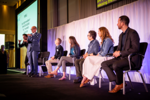 A panel discussion, featuring Dr Joseph Murray, Dr Katie Rogers, Tom Lichy, Tilly Hawkes and Dani Sive, is taking place on stage. Dr Joseph Murray is standing and signing, while an interpreter signs behind him, and the others watch and listen. All are dressed in business casual attire. A large screen behind them displays partially visible text with their names listed.